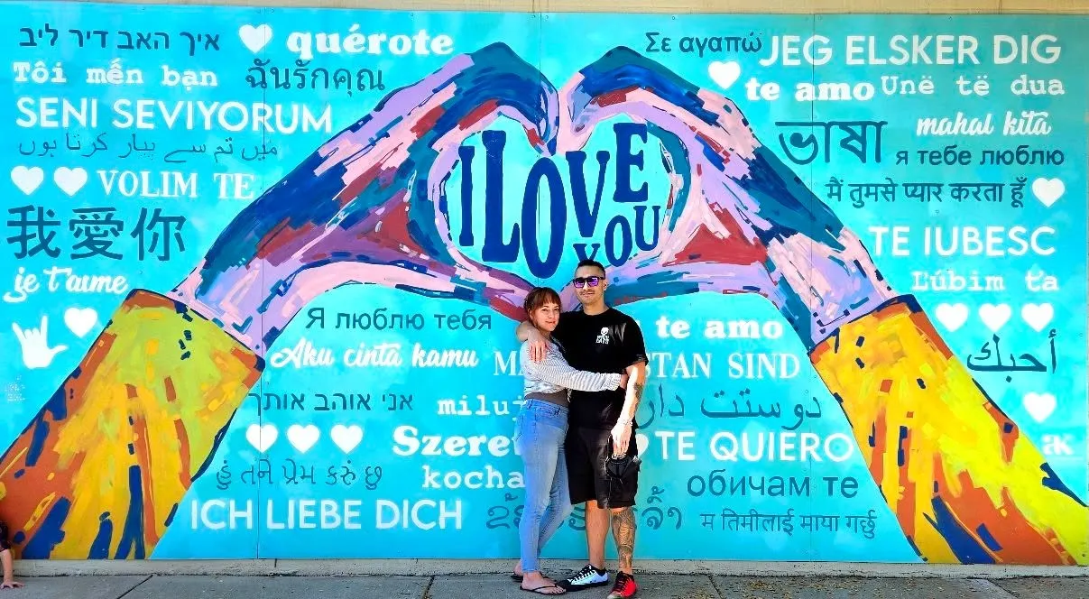Couple posing together smiling in front of a mural of two hands forming a heart with 'I Love You' written in many languages around them, Aiken, SC