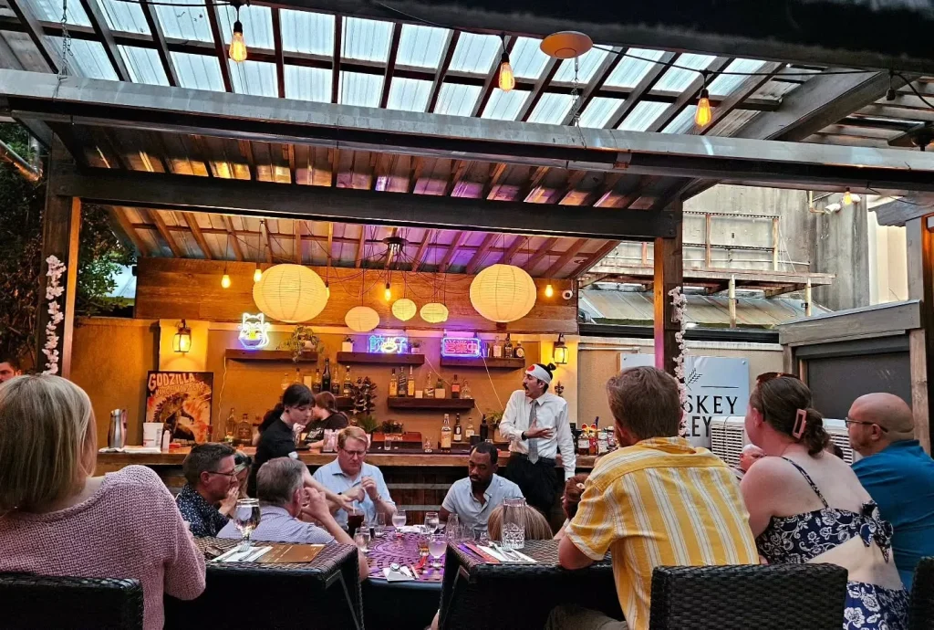 Outdoor covered patio bar/restaurant at dusk, groups of people seated at tables dining and socializing, string lights and paper lanterns, partial sign reading Whiskey Alley, Aiken, SC