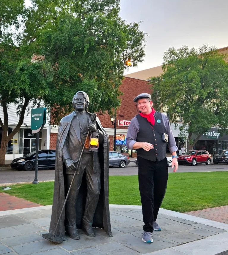Augusta Adventure Tours, Augusta, GA: a man in a period-style flat cap, vest, and red bandana posing beside a bronze statue of a man in a cloak holding a lantern, on a downtown street corner with storefronts and parked c