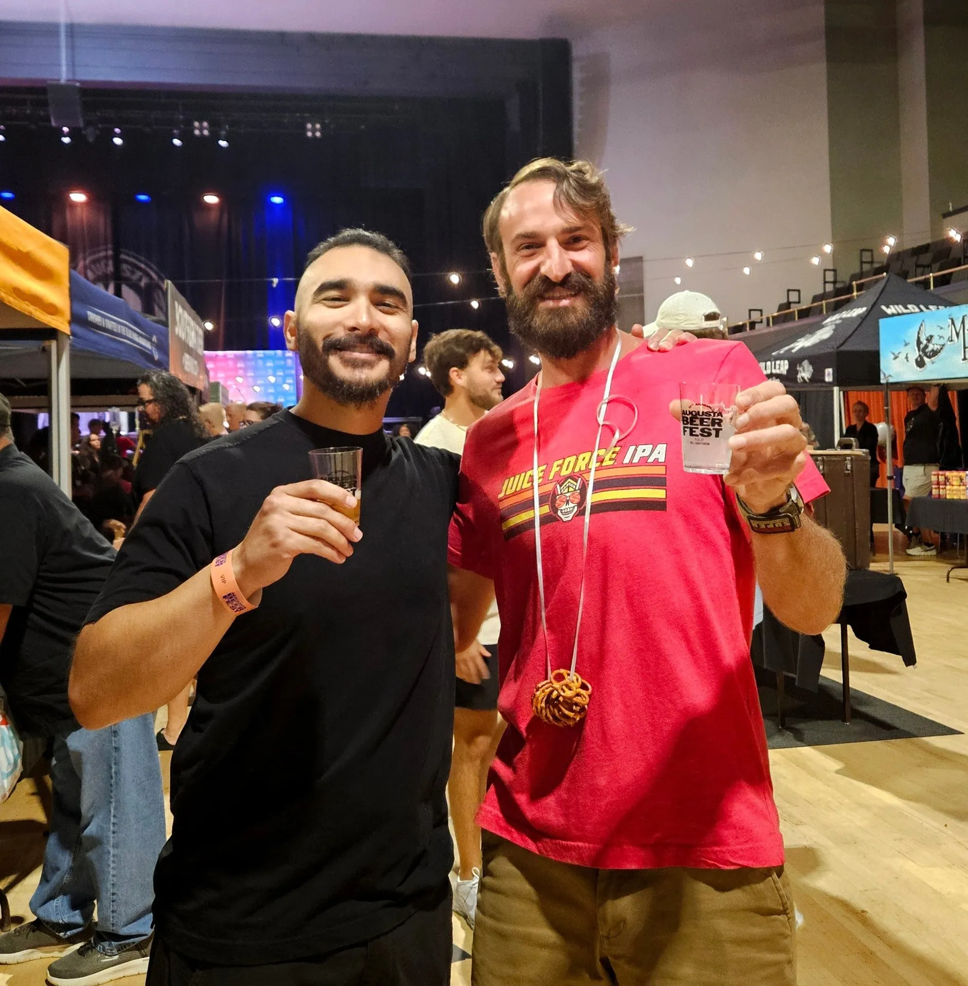 Augusta Beerfest, Augusta, GA: two smiling men at an indoor beer festival holding small beer tasting cups, crowd and vendor tents in background, one wearing a Juice Force
