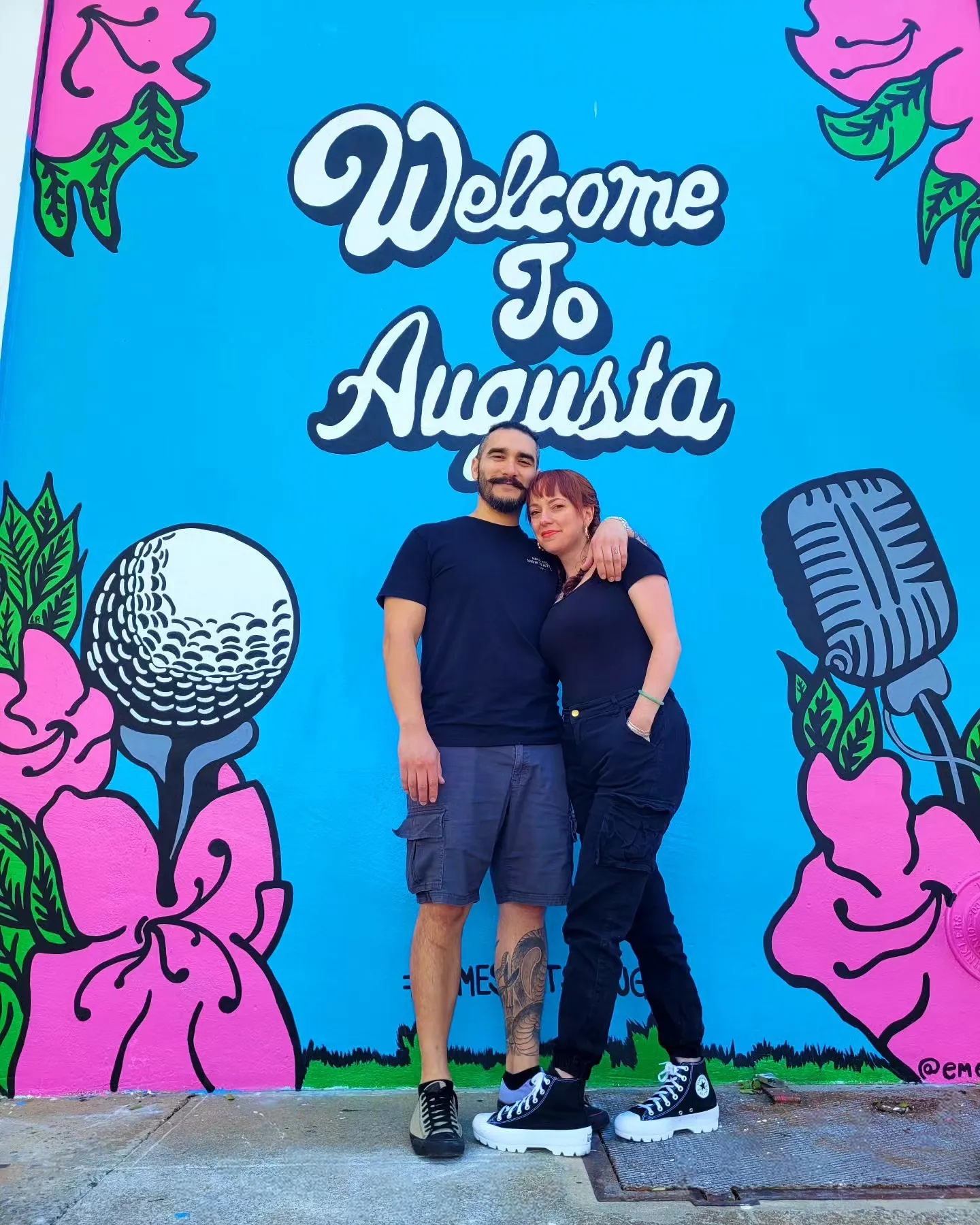 Couple posing arm in arm in front of a colorful mural reading Welcome To Augusta with golf ball, microphone, and pink floral graphics, Augusta, GA