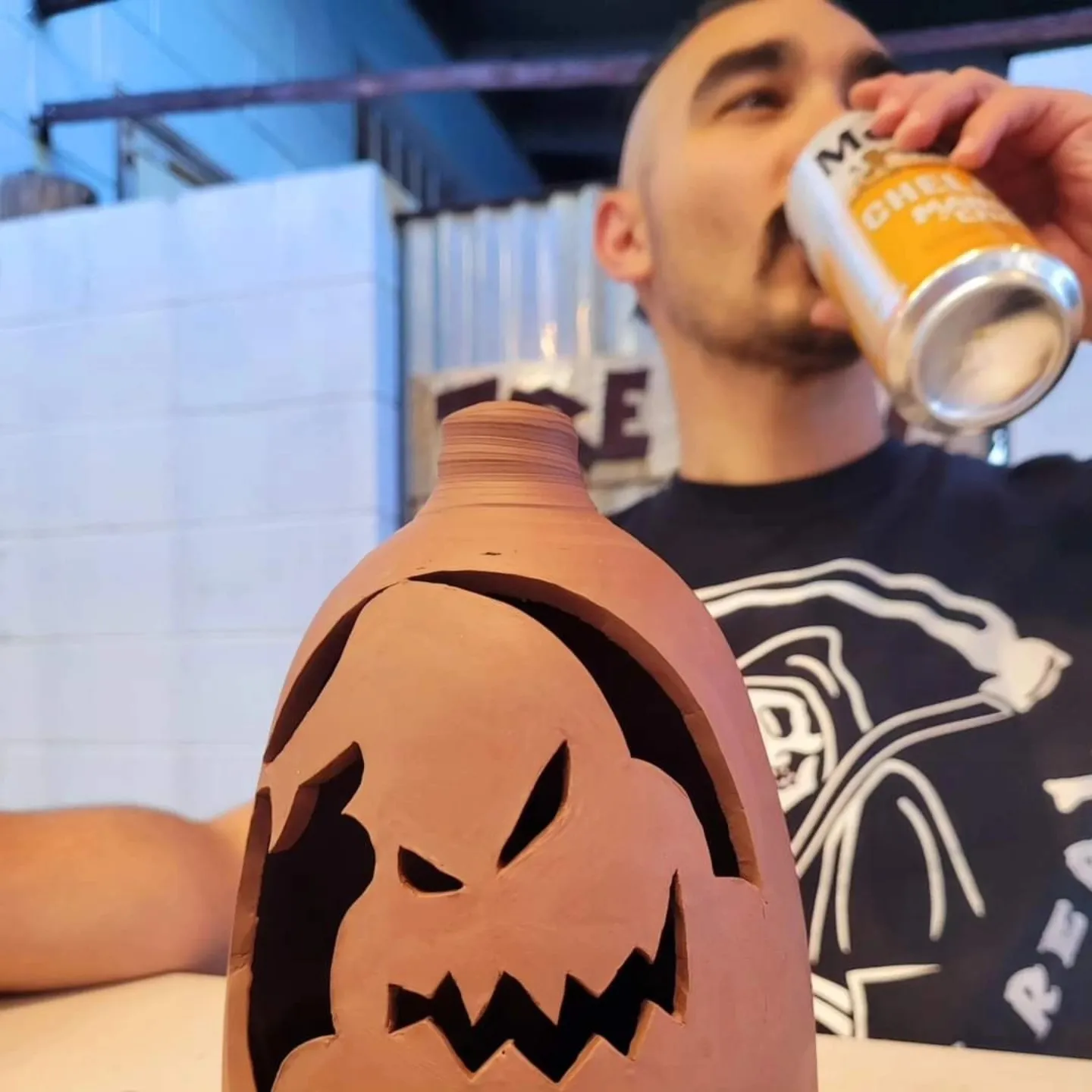 Man drinking from a beer can next to a carved unfired clay jack-o-lantern piece on a pottery studio table, industrial brick/cinderblock, Augusta, GA