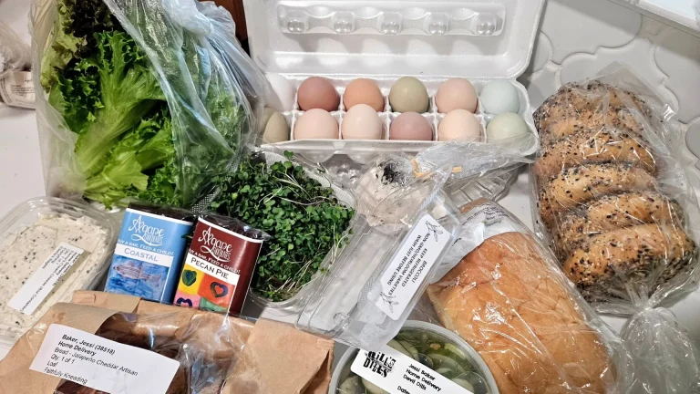 Augusta Locally Grown, Evans, GA: flat-lay of a local farm box: mixed lettuce greens, a carton of multicolor eggs, bagels, sliced bread, cheese wedge, honey packet, pecan pie mix, and a small salad container on a table