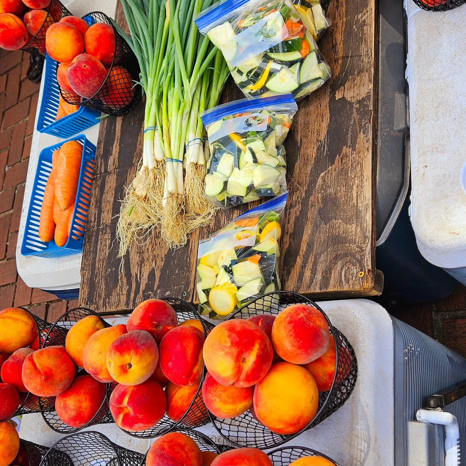 Overhead view of a farmers-market table loaded with fresh peaches in wire baskets, bundled green onions, carrots, and bagged pre-chopped vegetables, brick sidewalk in background, Augusta, GA