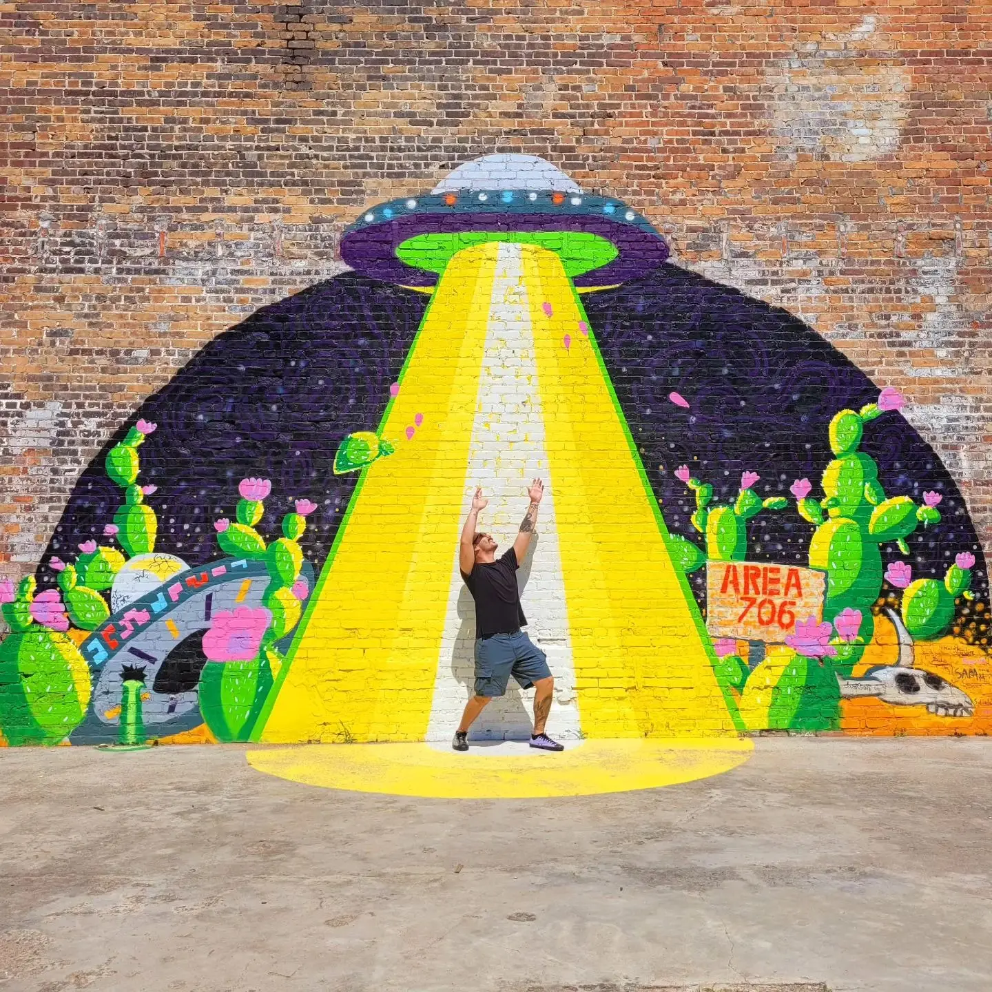 Colorful UFO abduction mural on brick wall with cacti and desert skull, text Area 706, person posing with arms raised standing in the painted beam of light, Augusta, GA