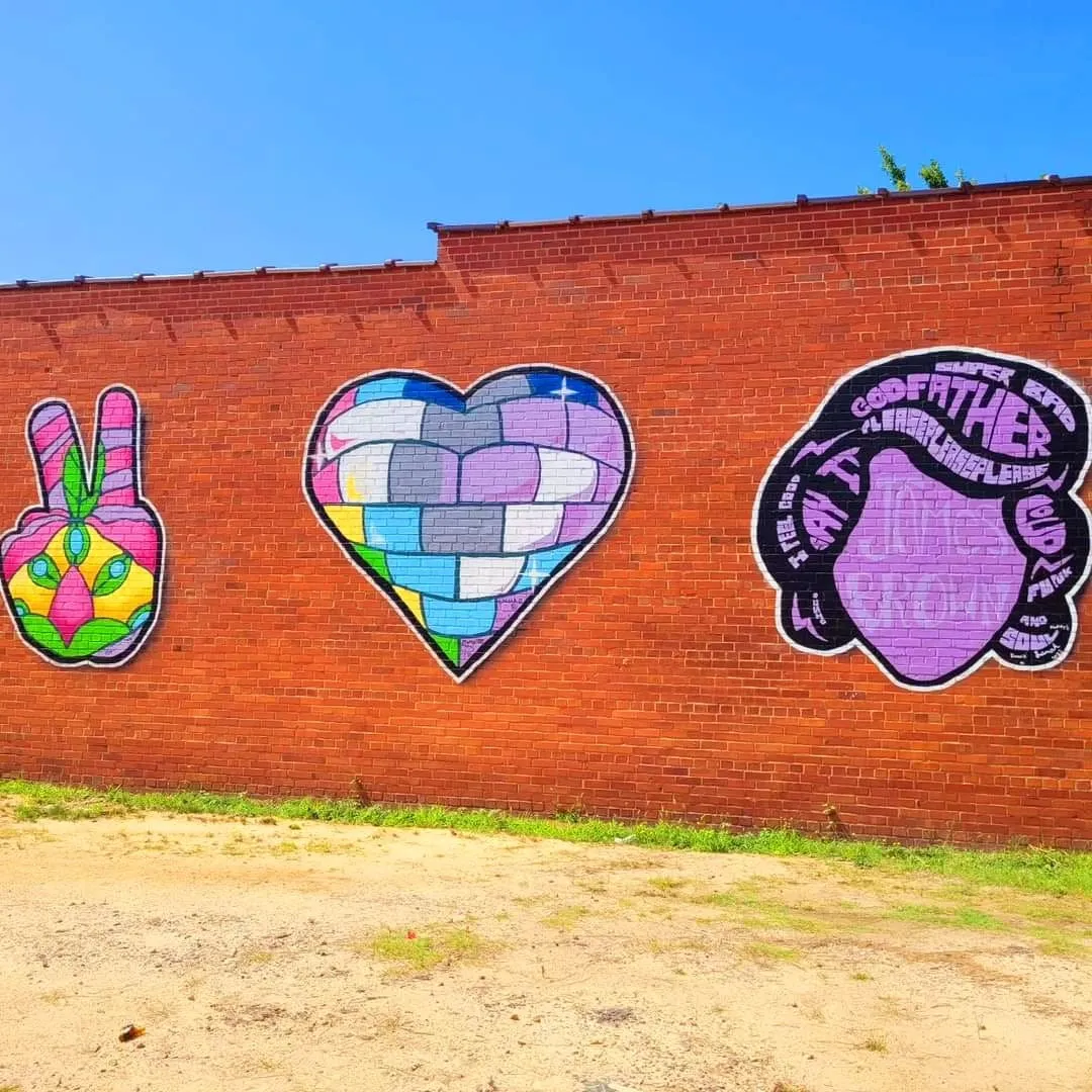 Three colorful murals painted on a red brick wall: a peace-sign hand, a patchwork heart, and a purple lettering design with a face silhouette, Augusta, GA