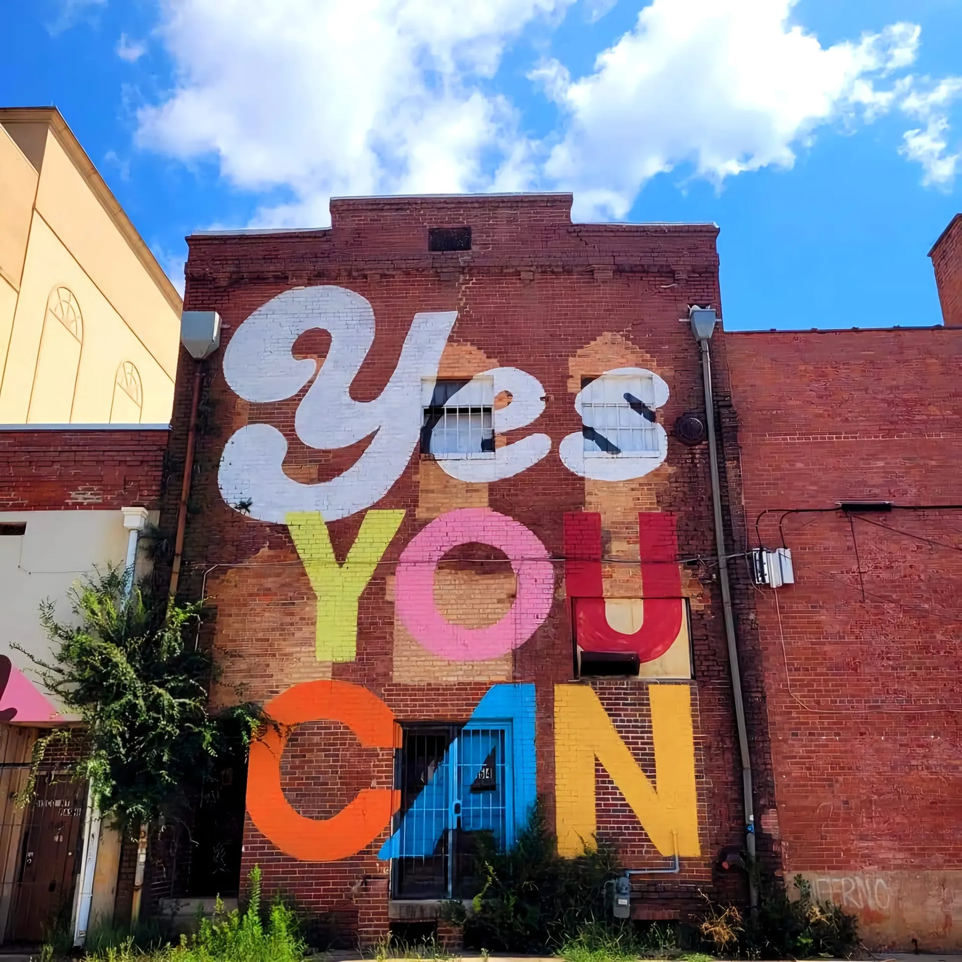 Colorful typography mural painted on brick building reading Yes You Can in large multicolor letters, blue sky with clouds, small alley, Augusta, GA