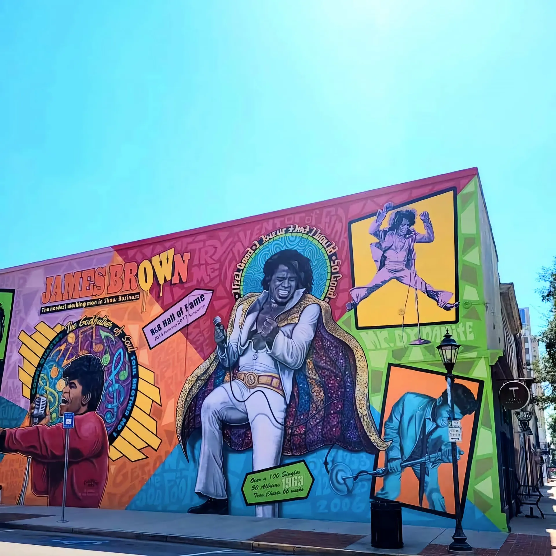 Large colorful mural of James Brown performing, multiple panels showing him singing and dancing, text James Brown and stats about hit singles, downtown street corner, Augusta, GA