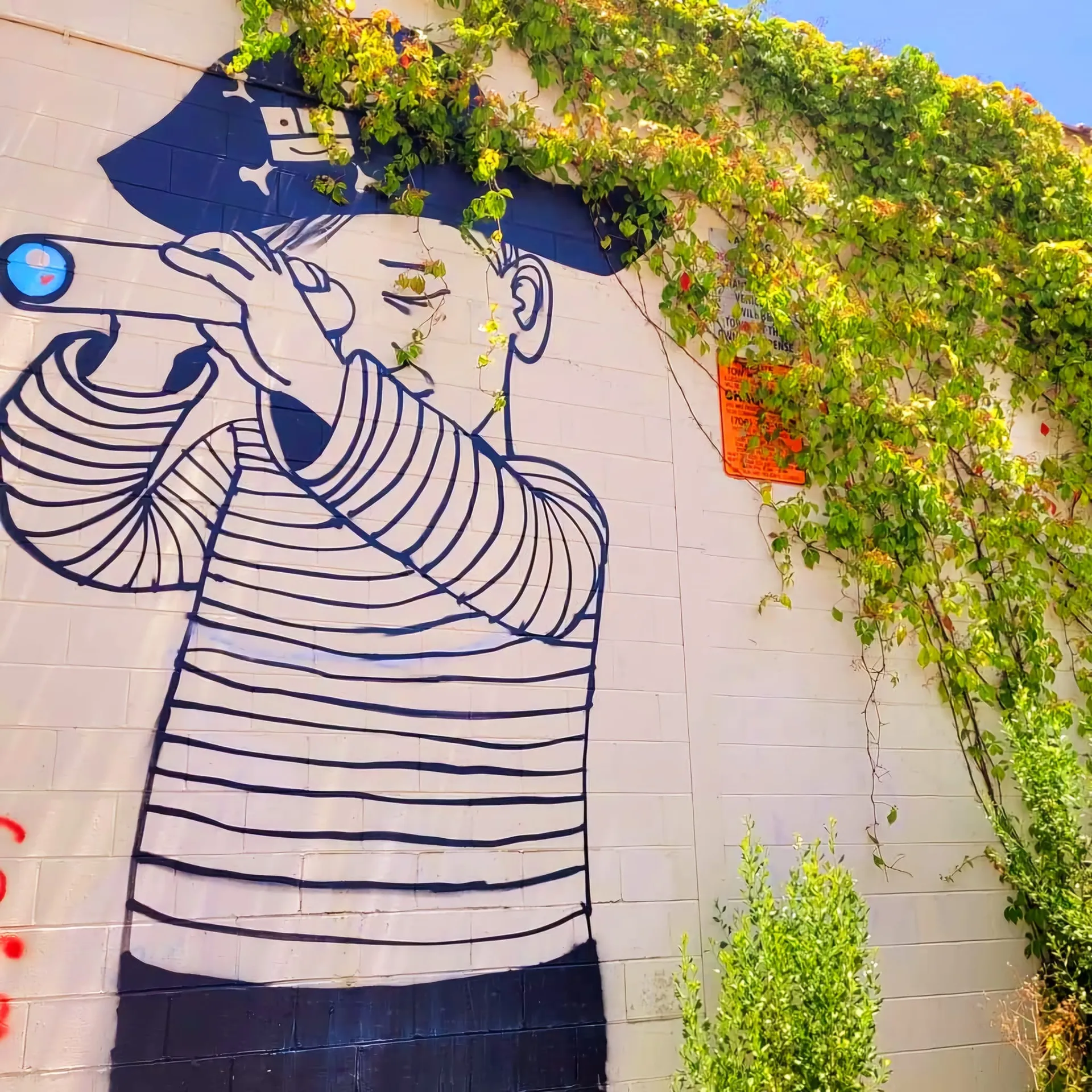 Building wall mural of a stylized sailor/fisherman figure in striped shirt and cap drinking from a bottle/horn, climbing vines and green foliage draping over the wall, Augusta, GA