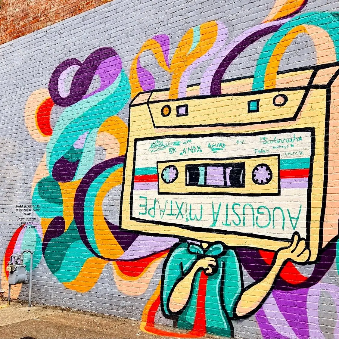 Colorful street mural on brick wall of a cartoon figure holding a giant cassette tape reading Augusta Mixtape, abstract swirl shapes in, Augusta, GA