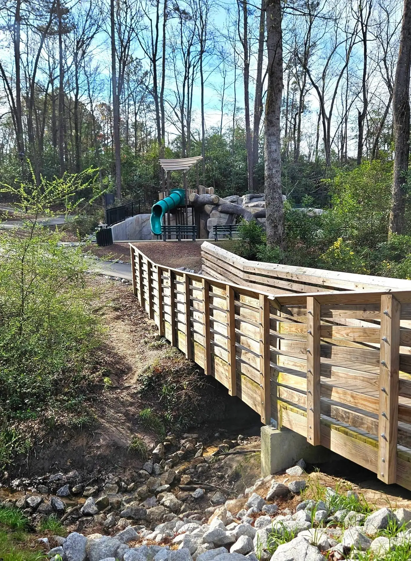 Blanchard Park, Martinez, GA: wooden boardwalk bridge over a rocky creek at a wooded park, with a playground slide and pavilion visible in the background