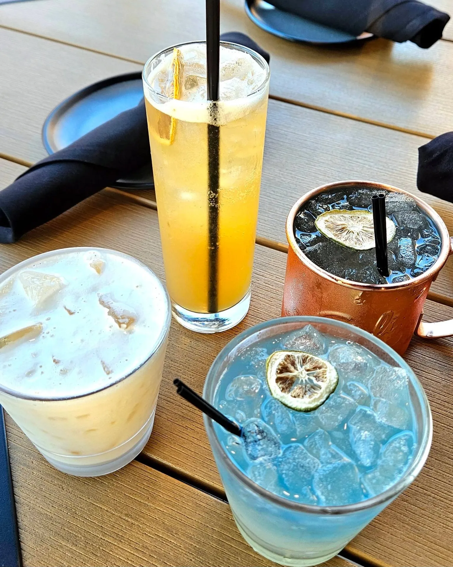 Brink's Tavern, North Augusta, SC: four cocktails on wooden tabletop: tall orange drink, cream-colored drink with ice, copper mug Moscow mule, blue cocktail with dried citrus wheel garnish
