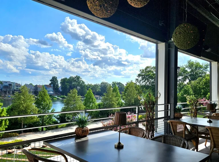Drift Raw Bar, North Augusta, SC: elevated open-air restaurant patio with wicker chairs and dark tables overlooking a river, bridge, and treeline, hanging woven pendant lights, blue sky with clouds