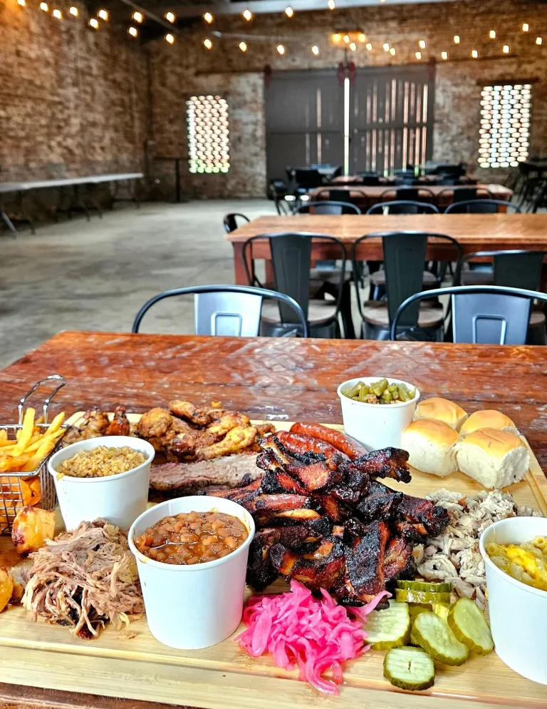 5th & Fire, Augusta, GA: large BBQ platter on a wooden board with brisket, ribs, pulled meat, baked beans, mac and cheese, coleslaw, pickled red onion, pickles, rolls, and fries, on a table inside a rustic exposed-brick