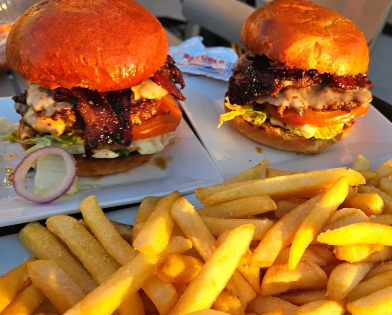 FreshTake Grocery, Augusta, GA: two burgers on toasted orange/red buns with bacon, lettuce, tomato and pickled onion, served on a white plate with a large pile of golden french fries in foreground, outdoor evening light