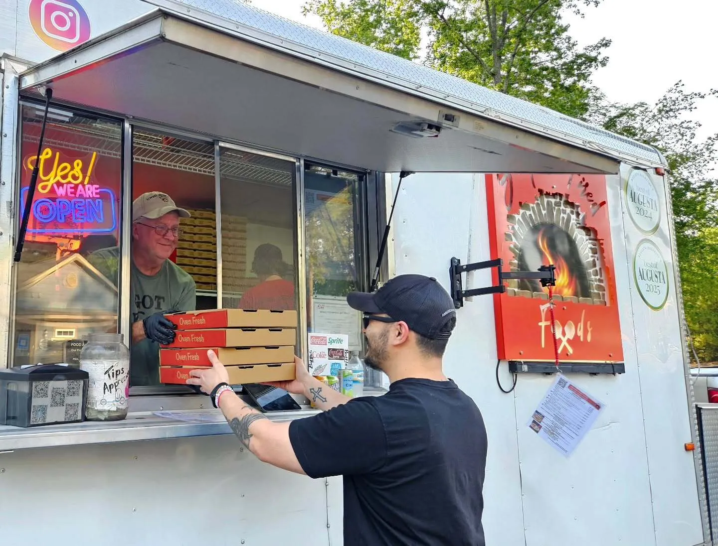 Wood-fired pizza food truck with neon Yes We Are Open sign, worker handing pizza boxes through the window to a customer, poster of pizza, Grovetown, GA