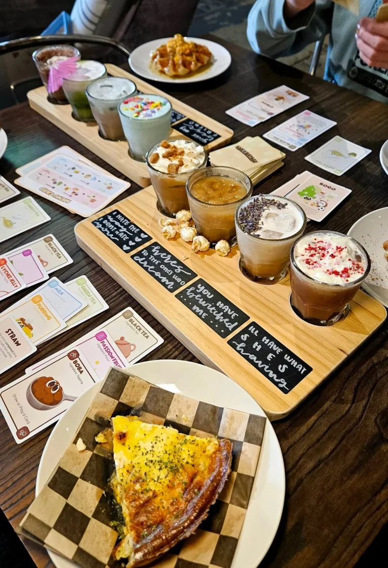 Harlem Java House, Harlem, GA: two wooden flight boards of specialty coffee drinks with chalkboard name tags, quiz/trivia cards scattered on table, a slice of quiche in foreground, waffle plate in background, people's ha