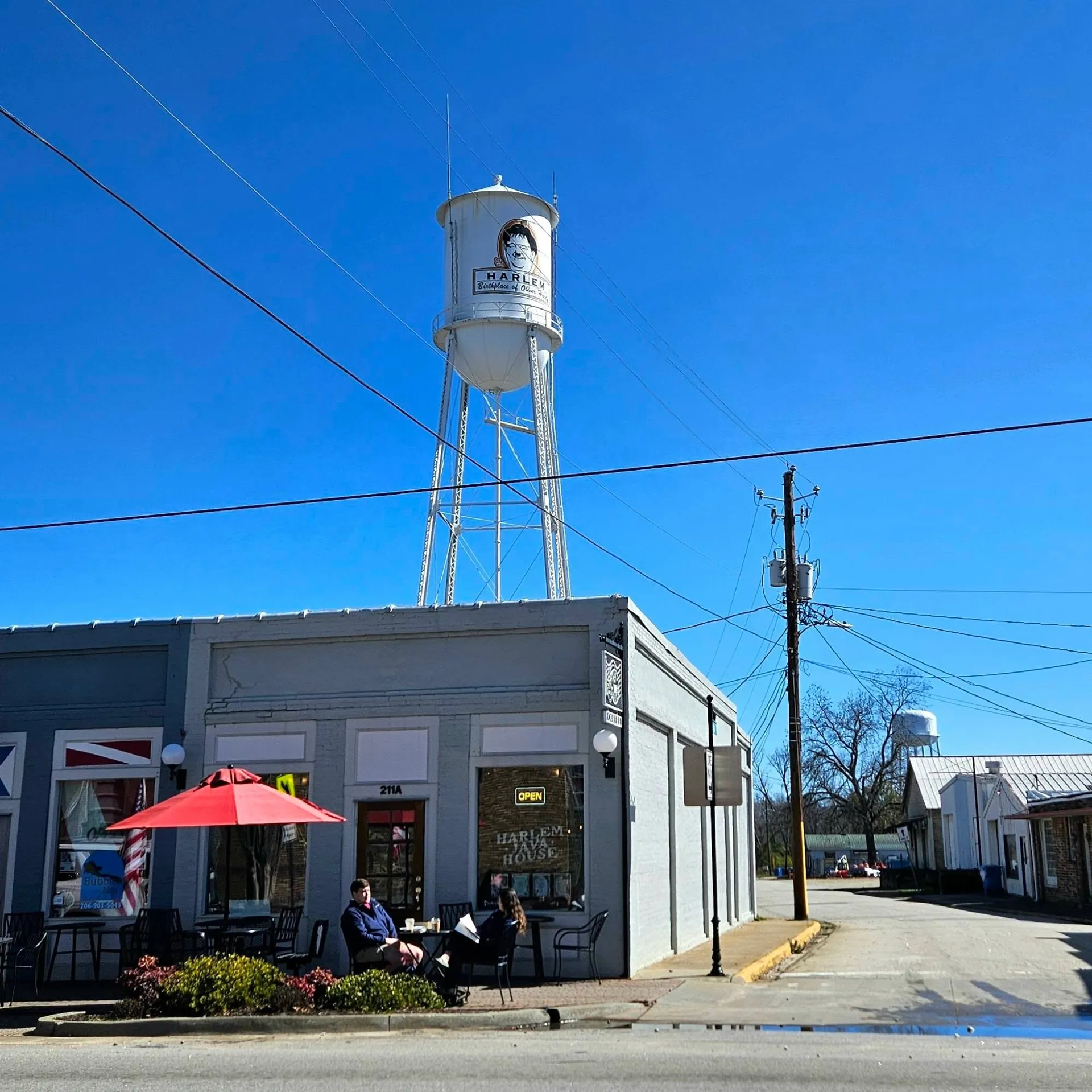 Harlem Java House, Harlem, GA: small-town street view with a water tower labeled Harlem behind a storefront cafe with a red patio umbrella and people seated outside under clear blue sky