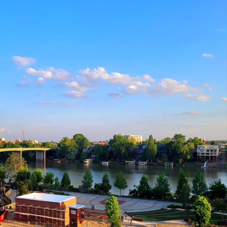 Jackson's Bluff, North Augusta, SC: elevated scenic view of a river with a bridge, tree-lined banks, riverside amphitheater/plaza, and city buildings in the distance under a blue sky with scattered clouds at golden hour