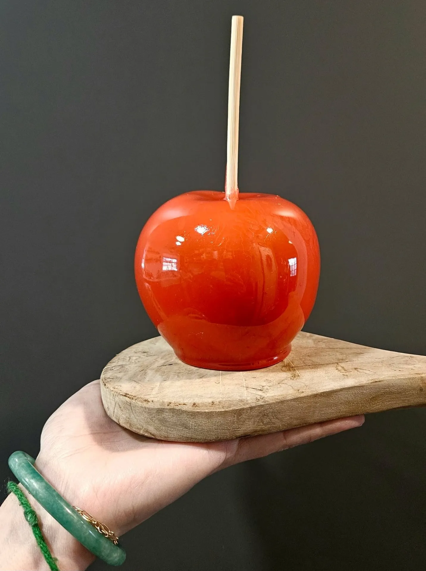 Kimmie's Kandy Koated Kreation, Augusta, GA: close-up of a glossy red candy apple on a wooden paddle, held on an open palm wearing a green bracelet, against a dark background