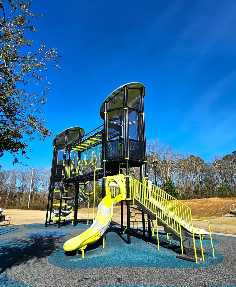 Lakeside Park, Evans, GA: modern playground with yellow and black slide and climbing structure on blue rubber surfacing, clear blue sky, trees in background