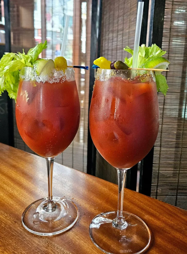 Manuel's Bread Cafe, North Augusta, SC: two Bloody Mary cocktails in stemmed glasses garnished with celery, olives, and pickle on a wooden bar counter, cafe interior with woven room divider