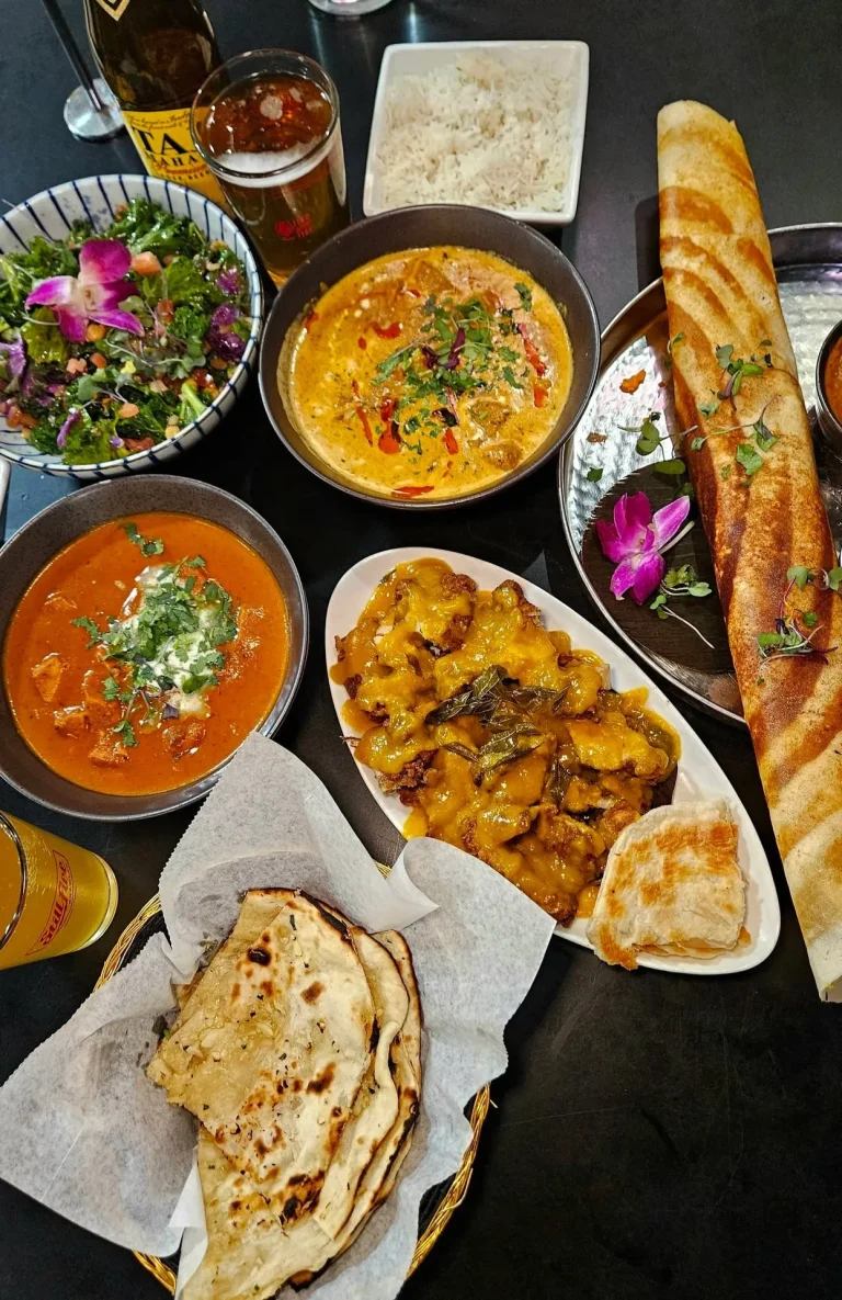 Namaste Indian Street Food, Augusta, GA: indian food spread on black table: orange curry, yellow korma-style curry, salad with flowers, white rice, stuffed naan/paratha bread, long crispy dosa roll, glass of beer