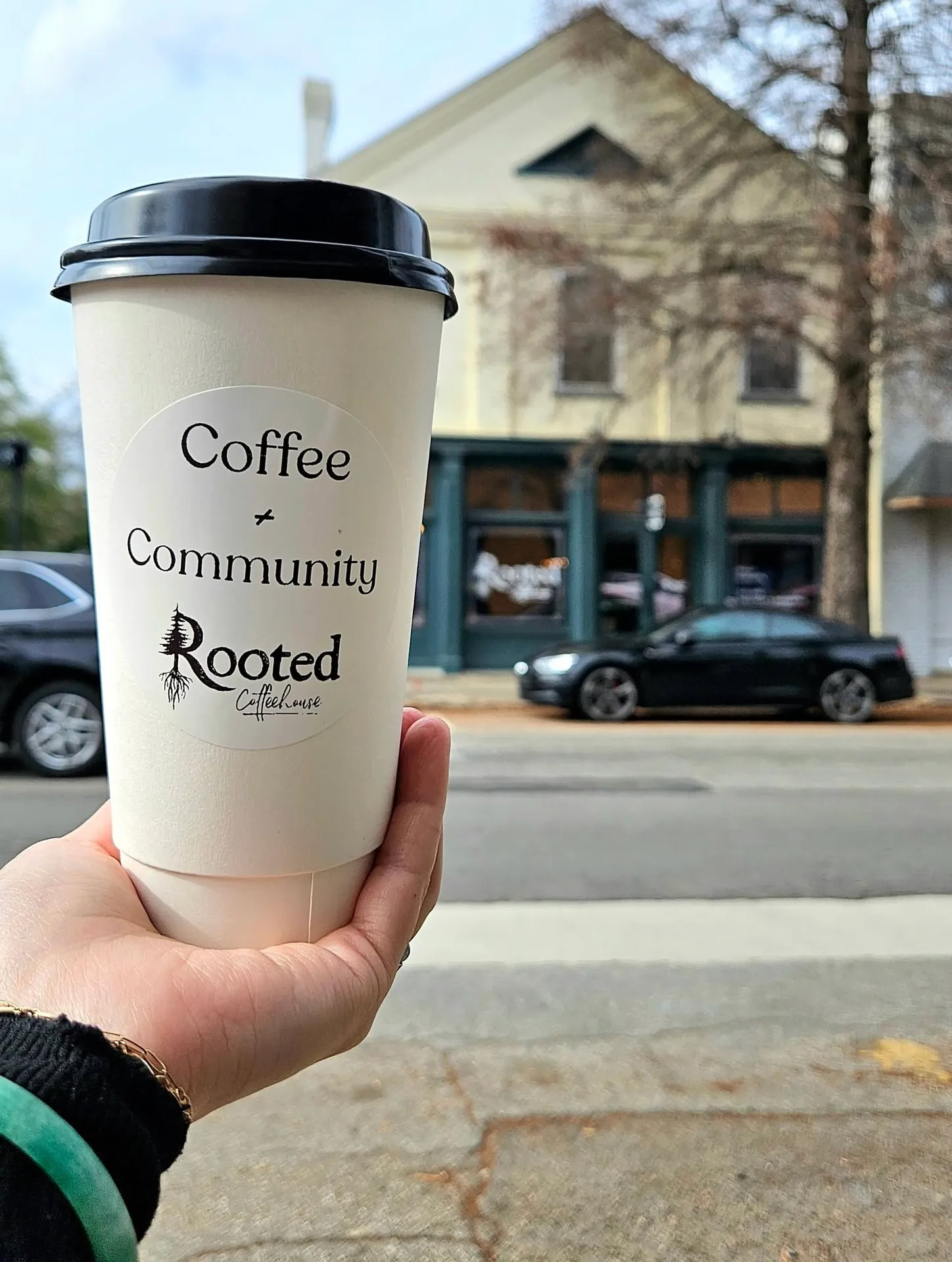 Rooted Coffeehouse, Augusta, GA: hand holding a white paper to-go coffee cup labeled 'Coffee + Community, Rooted Coffee House' with black lid, historic downtown street with parked cars and storefronts in background