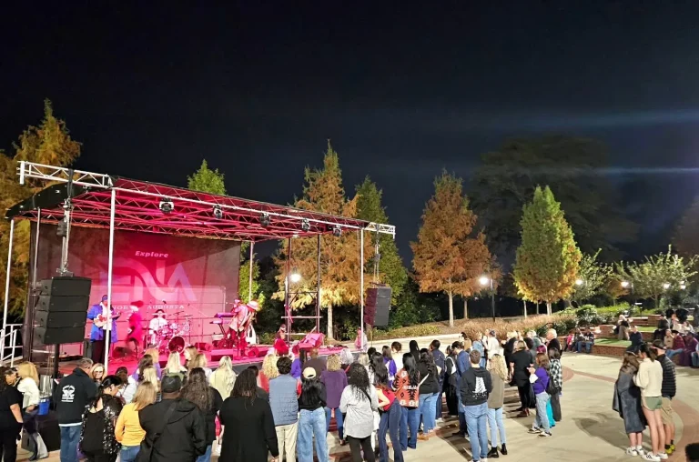 Sharon Jones Amphitheater, North Augusta, SC: nighttime outdoor concert with a band performing on a lit pink-hued stage under a truss rig, a crowd of spectators standing on the lawn watching