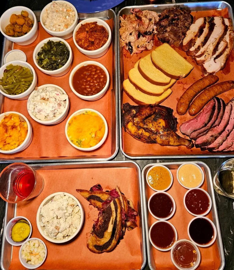 Smoke Barrels, Evans, GA: large BBQ feast spread on metal trays: sliced brisket, pulled pork, sausage links, cornbread, fried chicken, sides of mac salad, collard greens, baked beans, and an assortment of sauce cups