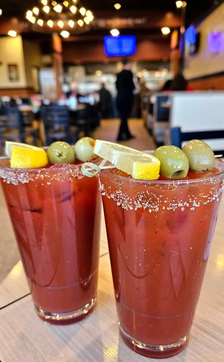 That Flippin Egg, Aiken, SC: two Bloody Mary cocktails with salt rims garnished with olives and lemon wedges, blurred restaurant interior with pendant lights and person in background