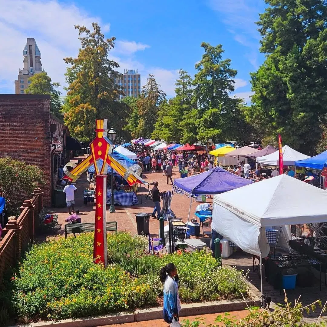 The, Augusta, GA: elevated view of an outdoor downtown market with white and colorful vendor tents, crowds walking, garden beds, and city skyline in