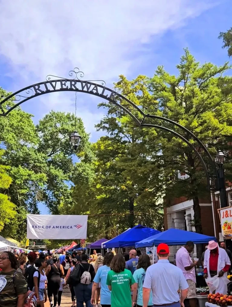 The, Augusta, GA: crowd of people walking under a metal Riverwalk arch sign at an outdoor market with blue vendor tents and a Bank of America banner