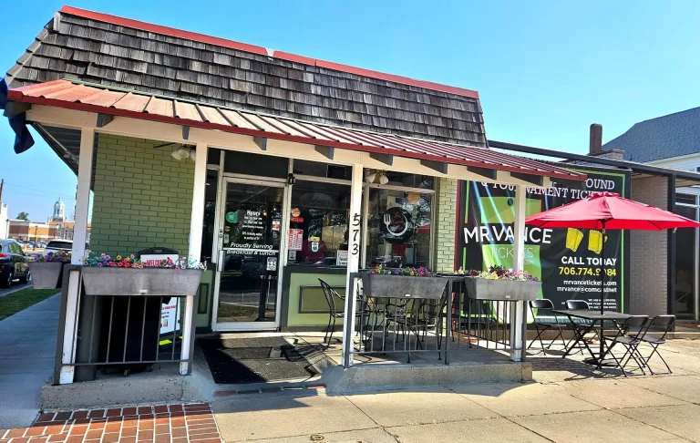 The Brunch House Of, Augusta, GA: storefront exterior of a small restaurant with green shingle siding, red-shingled awning roof, outdoor patio tables with a red umbrella, and address 5713 visible; daytime, clear sky