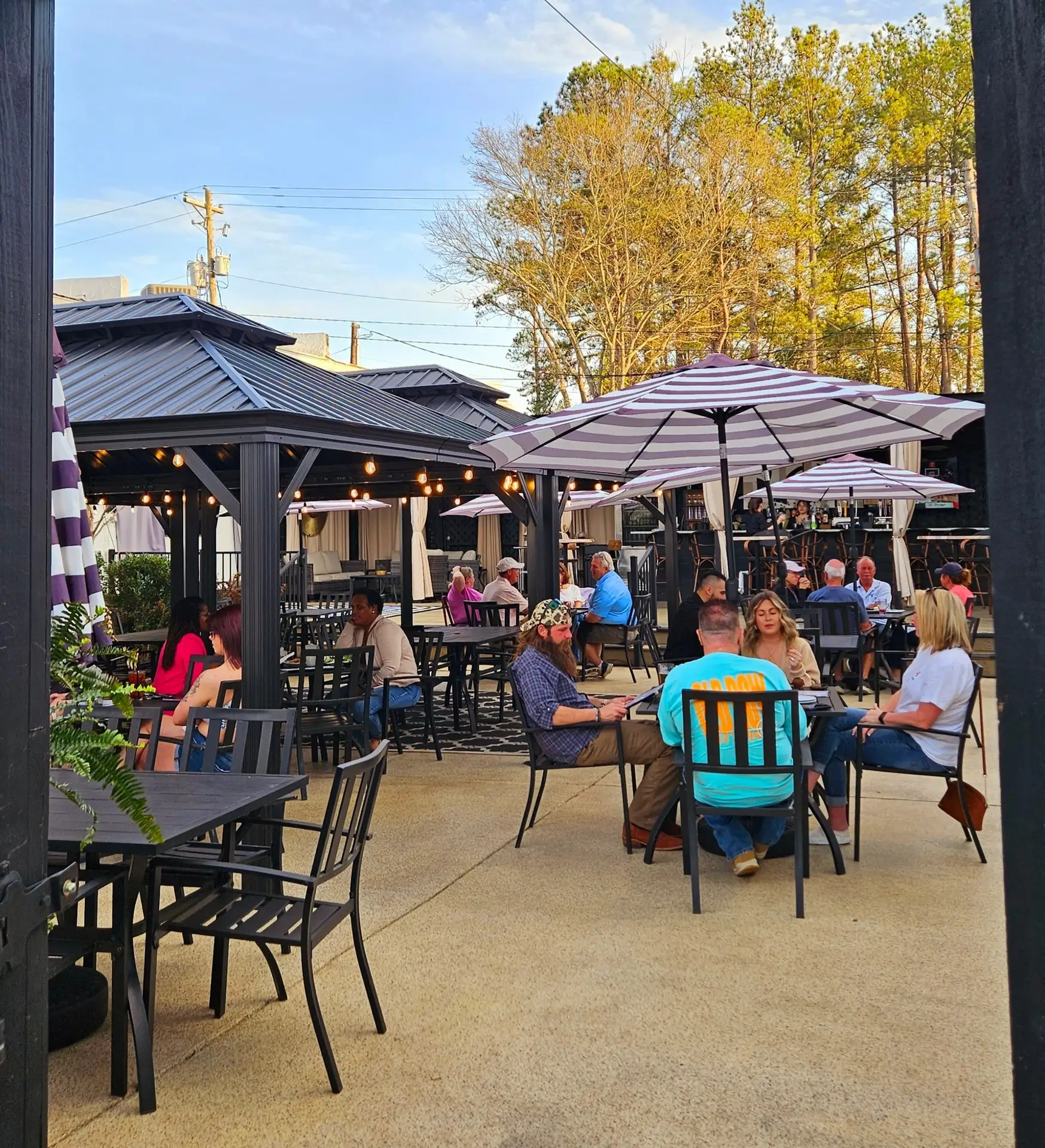 The Feed Sack, Aiken, SC: busy outdoor restaurant patio at dusk with striped umbrellas, string lights, a covered gazebo bar area, and multiple groups of people seated at tables