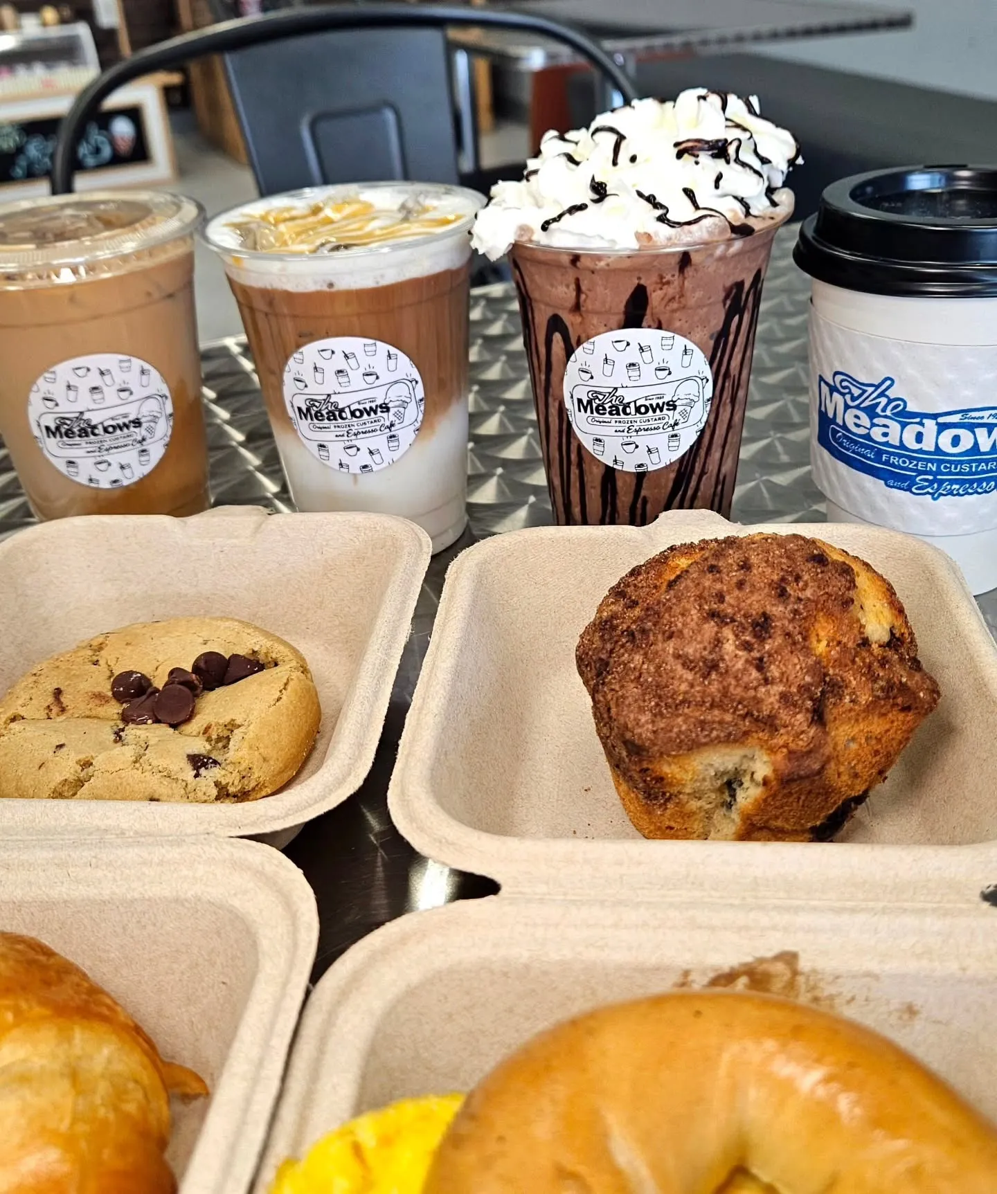 The Meadows, Grovetown, GA: bakery/cafe spread: iced coffee, iced latte, whipped-cream-topped chocolate frozen custard cup, and a to-go coffee cup labeled The Meadows, with pastries in trays below - chocolate chip cookie