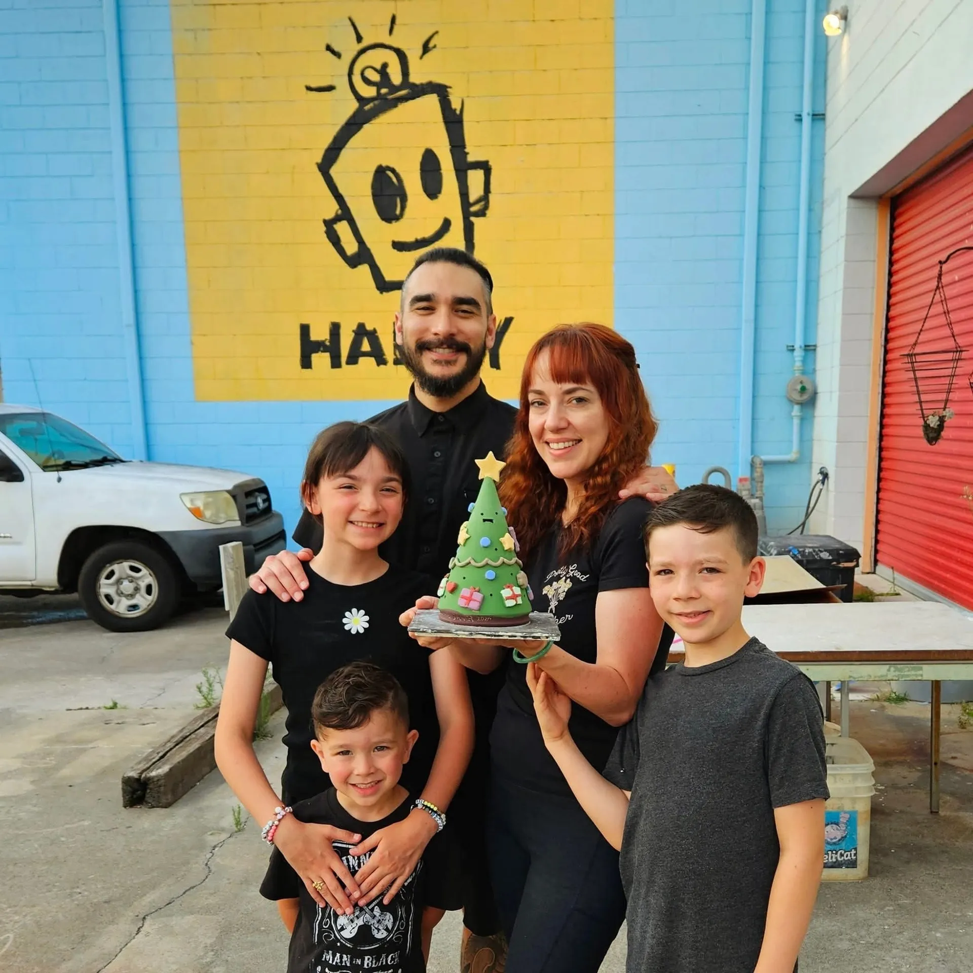 Tire City Potters, Augusta, GA: a family of five (two adults, three children) posing outdoors in front of a yellow mural with a smiling robot cartoon and HAPPY text, holding a hand-painted ceramic Christmas tree figurine
